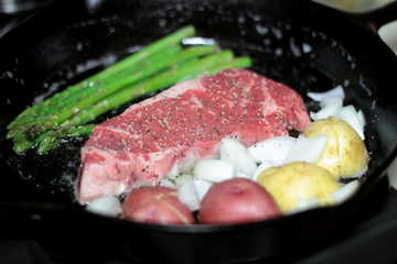 New York strip steak frying in a cast iron pan with potatoes and asparagus on a natural gas stove top.