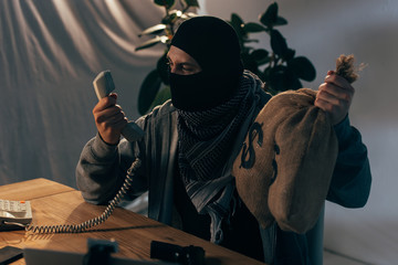 Angry terrorist in mask holding money bags and looking at handset