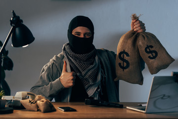 Terrorist in mask holding bags with dollars and showing thumb up