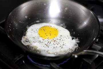 Egg sunny side up with salt and pepper, frying in a pan on the stove.