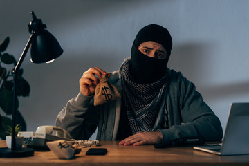 Sad criminal in black mask sitting at table and holding small bag with dollars