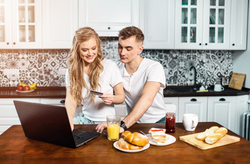 Obraz na płótnie Canvas Young happy couple sitting at the kitchen looking at laptop and holding credit card