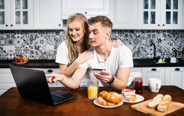 Excited young couple shopping online, holding credit card and looking at laptop while sitting at the kitchen