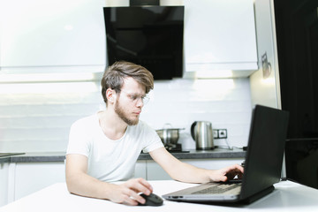 bearded young man wearing glasses in the kitchen using a laptop, searching the Internet for furniture and home appliances