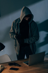 Criminal in mask and hoodie looking at laptop in dark room