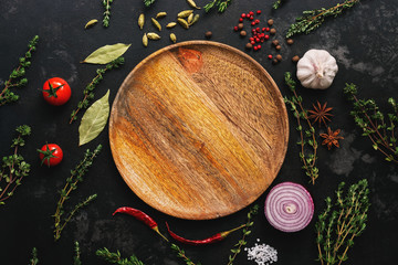 Obraz na płótnie Canvas Empty wooden plate and frame of spices, herbs and vegetables on a dark stone background. Top view, flat lay.