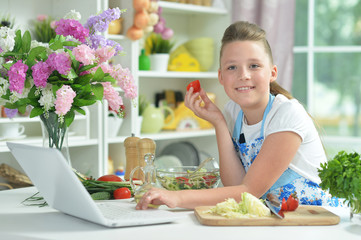 Portrait of cute teen girl using laptop while preparing fresh salad on kitchen table