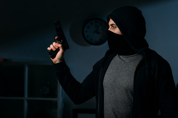 Burglar in mask holding pistol in dark room and looking away