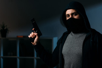 Robber in mask holding gun and looking at camera in dark room