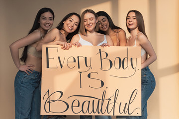five cheerful multicultural girls holding placard with "every body is perfect" inscription