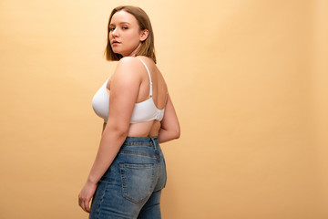 pretty overweight girl in blue jeans and bra looking at camera isolated on beige, body positivity...