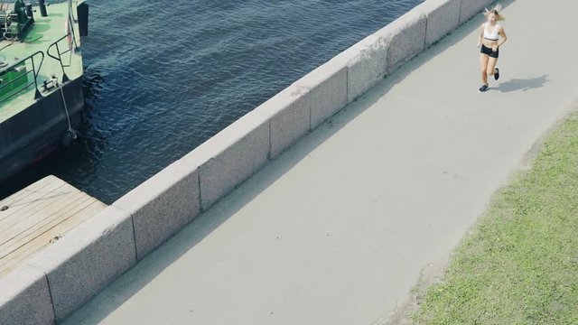 High angle of female athlete running along embankment next to river, stopping to check distance and speed on her smartwatch and proceeding marathon training
