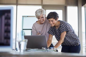 Two smiling businesswomen sharing laptop at desk in office
