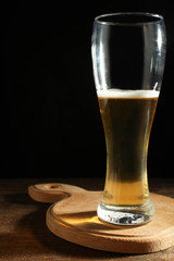 Light beer in glass goblet on wooden cutting board on black background in  dark. Concept alcohol.