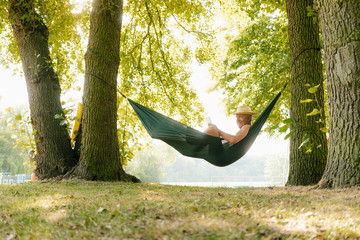 Senior man wearing straw hat relaxing in hammock at lakeshore reading book
