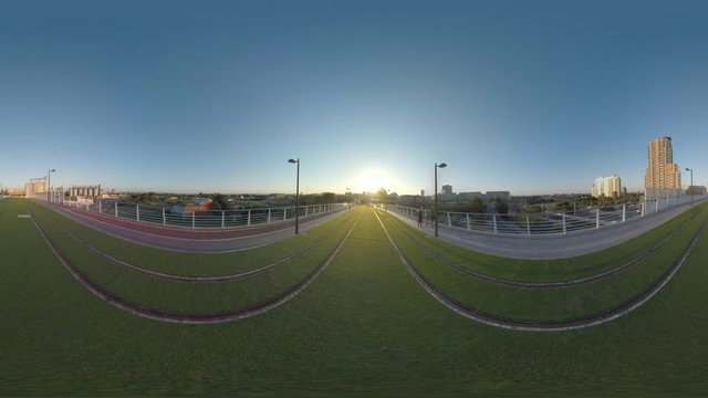 VALENCIA, SPAIN - JULY 15 2016: 360 VR video. View to the city at sunset from bridge with green lawn and rails. Family (with model release) walking alongside, some people having evening jogging there