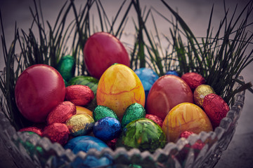 Bunter Ostereier und Schoko Eier in einer Kristallschüssel mit grünem Gras und dunkler Vignette als Hintergrund.