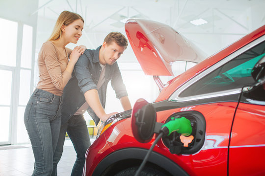 Young Family Buying First Electric Car In The Showroom. Attractive Couple Looking Under Eco-friendly Car Hood. Electric Car Sale Concept