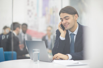 Fototapeta premium Portrait of young Middle-Eastern businessman speaking by phone and using laptop while working in office, shot from behind glass, copy space