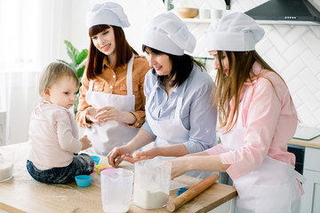 Cute little girl sitting on the table and her beautiful mother, aunt and grandmother in aprons are preparing dough for muffins and smiling while baking in kitchen at home. Mothers Day concept