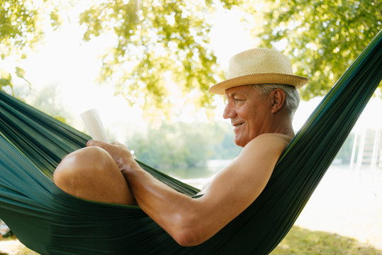 Senior man wearing straw hat relaxing in hammock at lakeshore reading book