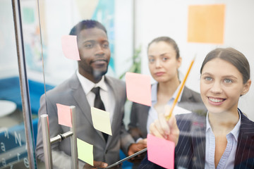 Multi-ethnic group of business people planning startup project placing sticky notes on glass wall,...