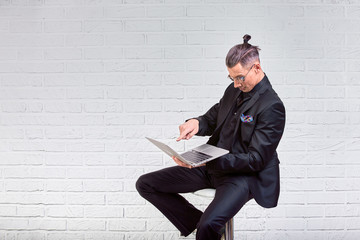 Handsome business man in glasses and suit holding laptop in hands and writing something. Side view. On white brick background