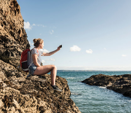 Young woman sitting on a rocky beach, taking pictures with her smartphone