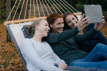 Happy mother with two teenage girls lying in hammock in garden in autumn using tablet