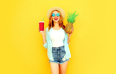 Portrait happy smiling woman holding pineapple, cup of juice having fun in summer straw hat, sunglasses, shorts on colorful yellow background
