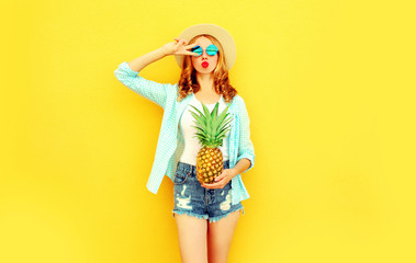 cool girl with pineapple blowing red lips sending sweet air kiss in summer straw hat, sunglasses, shorts on colorful yellow background