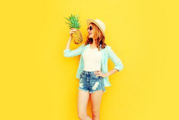 Portrait happy smiling woman holding pineapple having fun in summer straw hat, sunglasses, shorts on colorful yellow background