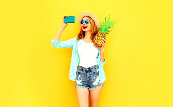 happy smiling woman with pineapple taking selfie picture by phone in summer round hat, sunglasses, shorts on colorful yellow background