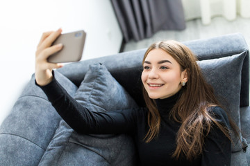 Happy emotional woman making funny selfie on smartphone while lying on sofa