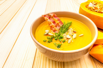 Homemade autumn hot pumpkin cream soup with smocked bacon and seeds. Raw pumpkin, fresh chives, dill, salt in spoon on wooden background, close up.
