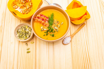 Homemade autumn hot pumpkin cream soup with smocked bacon and seeds. Raw pumpkin, fresh chives, dill, salt in spoon on wooden background.