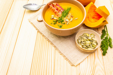 Homemade autumn hot pumpkin cream soup with smocked bacon and seeds. Raw pumpkin, fresh chives, dill, salt in spoon, vintage linen cloth on wooden background.