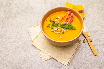 Homemade autumn hot pumpkin cream soup with smocked bacon and seeds. Raw pumpkin, fresh chives, dill, salt in spoon, vintage linen cloth on stone background close up.