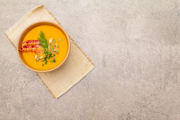 Homemade autumn hot pumpkin cream soup with smocked bacon and seeds. Raw pumpkin, fresh chives, dill, vintage linen cloth on stone background, top view.