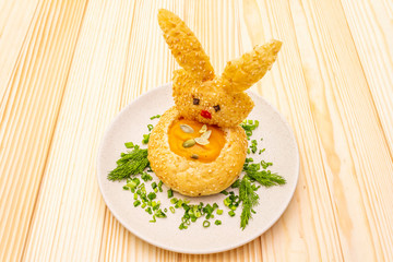 Easter bunny kids food concept. Warm pumpkin cream soup in fresh bun (roll) with herbs and pumpkin seeds in plate. Wooden background.
