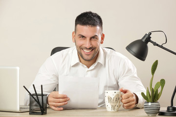 Portrait of a young cheerful businessman having a cup of coffee at his working desk, looking at the camera and smiling. Just an ordinary office day.