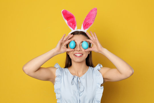 Beautiful woman in bunny ears headband holding Easter eggs near eyes on color background