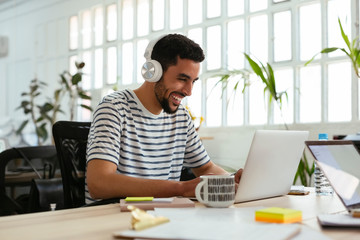 Laughing young man wearing headphones using laptop at desk in office