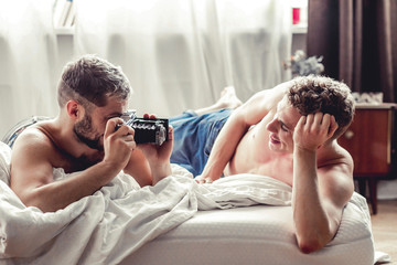 Gay man taking picture of his partner with old-fashioned camera in bed