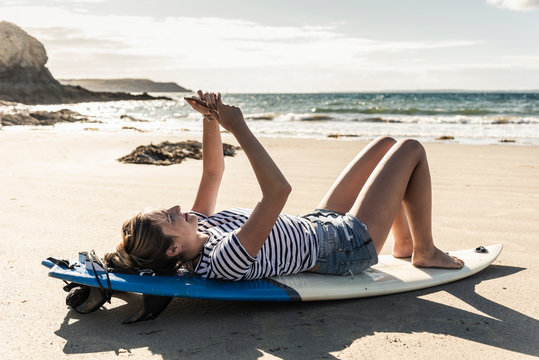 Young woman on the beach, relaxing on surfboard, using smartphone