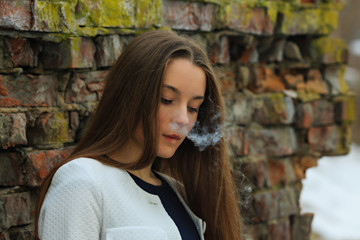 Vape teenager. Young pretty white girl in white cardigan smoking an electronic cigarette opposite destroyed brick wall on the street in the spring. Bad habit.