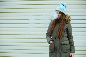 Vape teenager. Young pretty white girl in blue cap and dark green jacket smoking an electronic cigarette opposite modern brown background on the street in the winter. Bad habit.