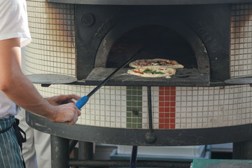 Pizza chef with blue and white apron placing two pizzas ( margarita and meat lovers) in a outdoor woodfired pizza oven.