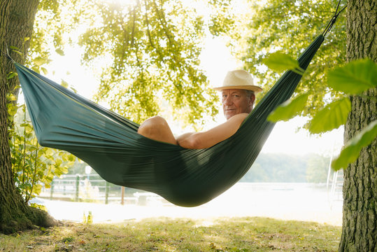 Senior man wearing straw hat relaxing in hammock at lakeshore