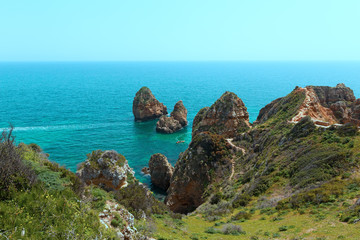 Pedestrian trails between cliffs with scenic views on turquoise sea waters and coastal line. Tourists explore grottoes on motorboat in Algarve, Portugal.